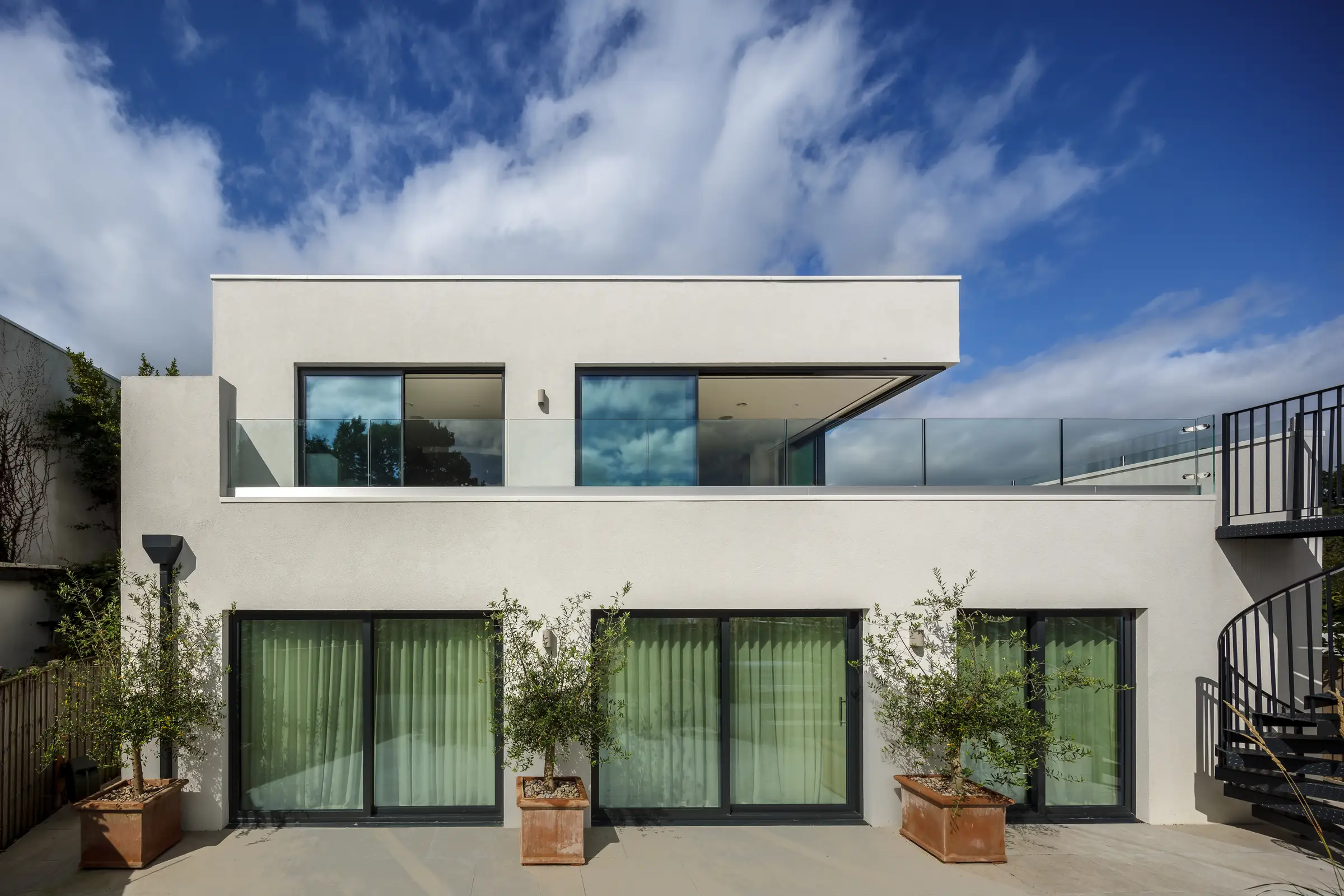 The rear elevation of Skippers Retreat — white render, full-height glazing, olive trees in terracotta pots and the black spiral staircase to the terrace