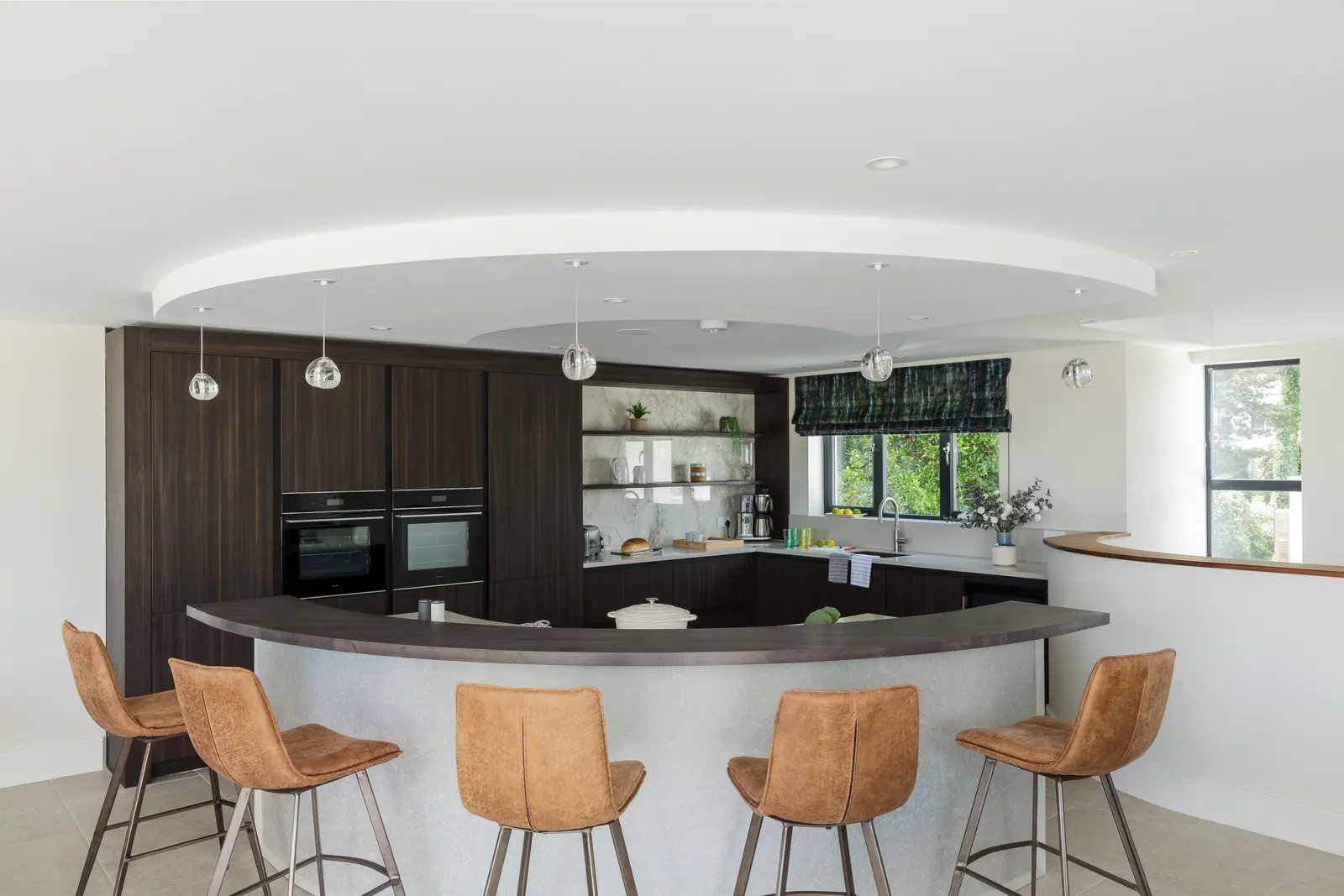 The kitchen — a curved island with tan leather bar stools, dark timber units and double electric ovens
